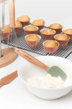 Kiddikutter sleikja og þeytari við bakstur, nýbakaðar muffins á grind í bakgrunni.