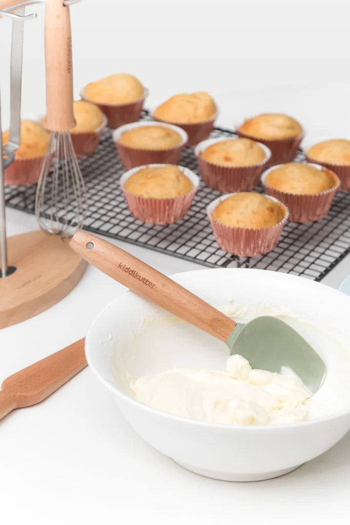 Kiddikutter sleikja og þeytari við bakstur, nýbakaðar muffins á grind í bakgrunni.