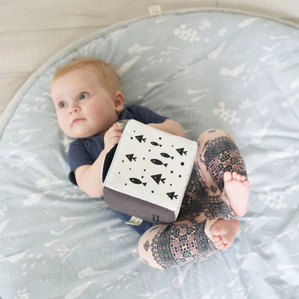 Baby lying on a mat holding a book with black fish illustrations