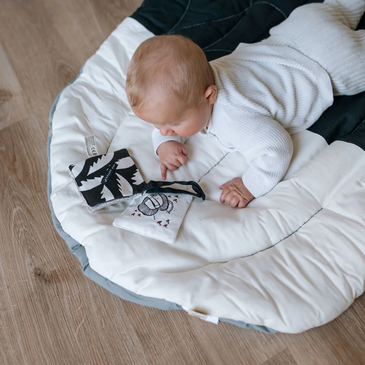 Baby lying on a white cushion with a black and white toy, sitting on a wooden floor.
